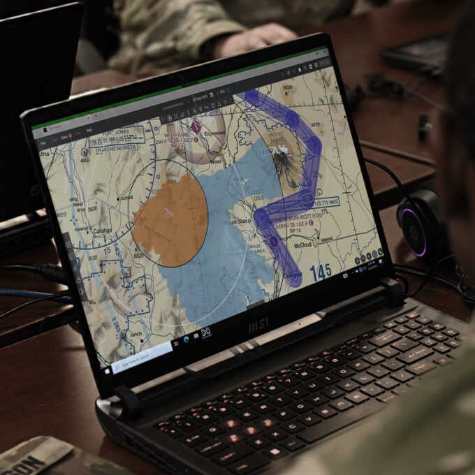 Soldier examining a laptop displaying a tactical map with blue routes and orange circle overlays on a desk.