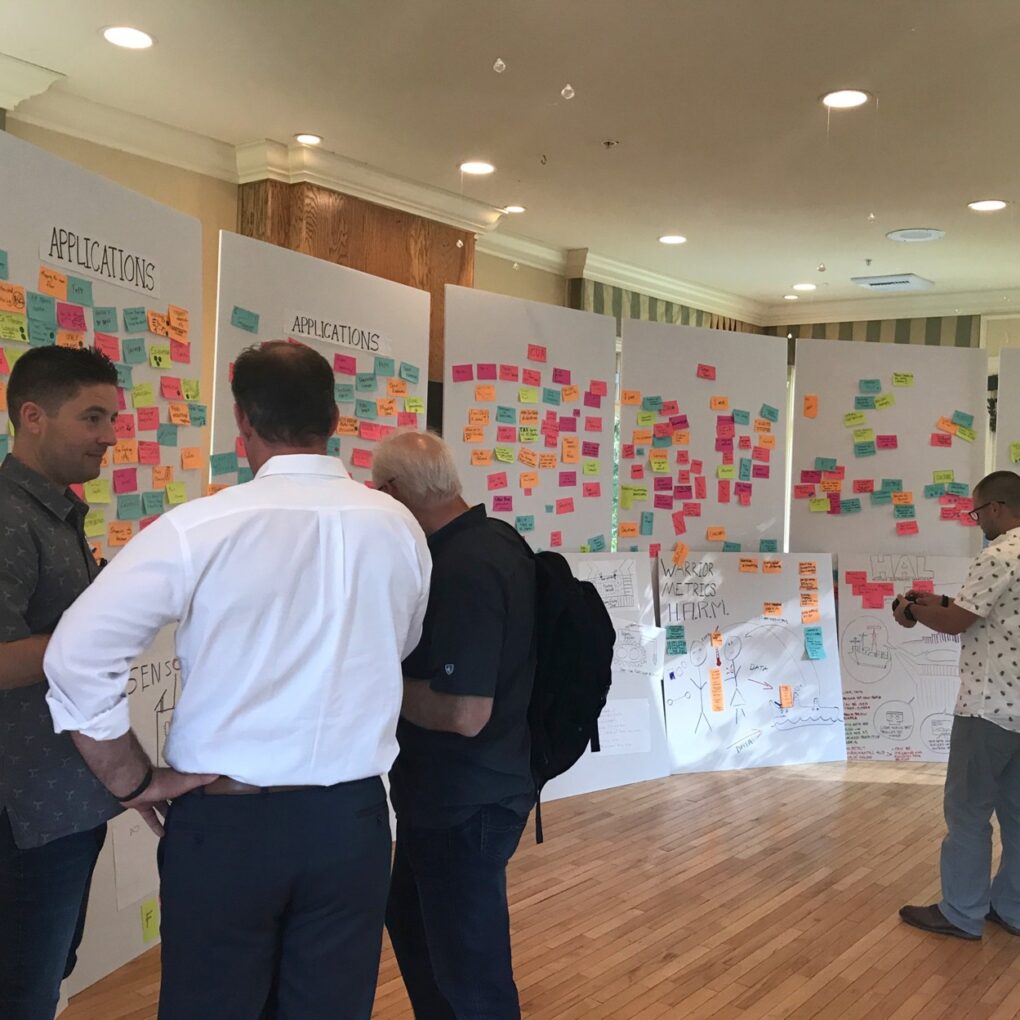 Group of people standing and looking at large white boards covered with multicolored sticky notes during a brainstorming session in a bright room.