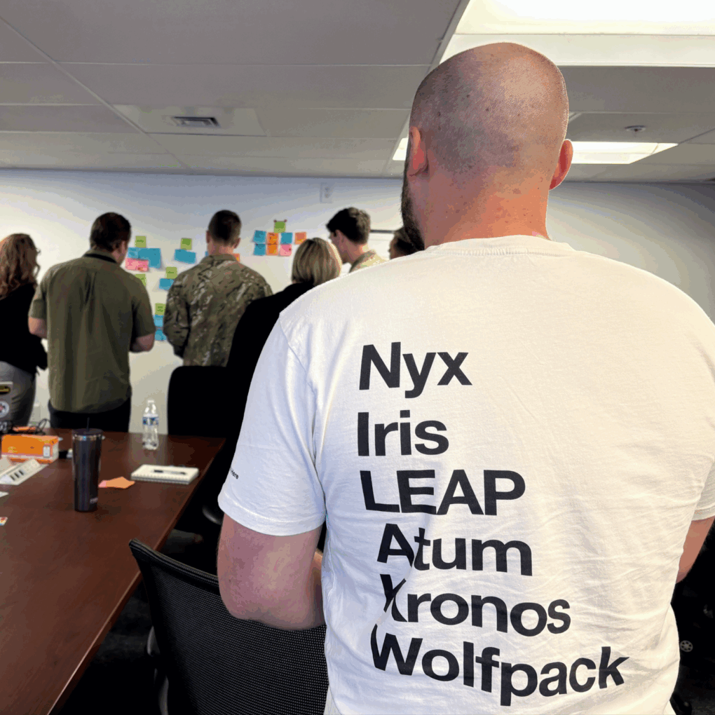 Back of a person in a white T-shirt with large black vertical text, facing a wall of colorful sticky notes in a meeting room.